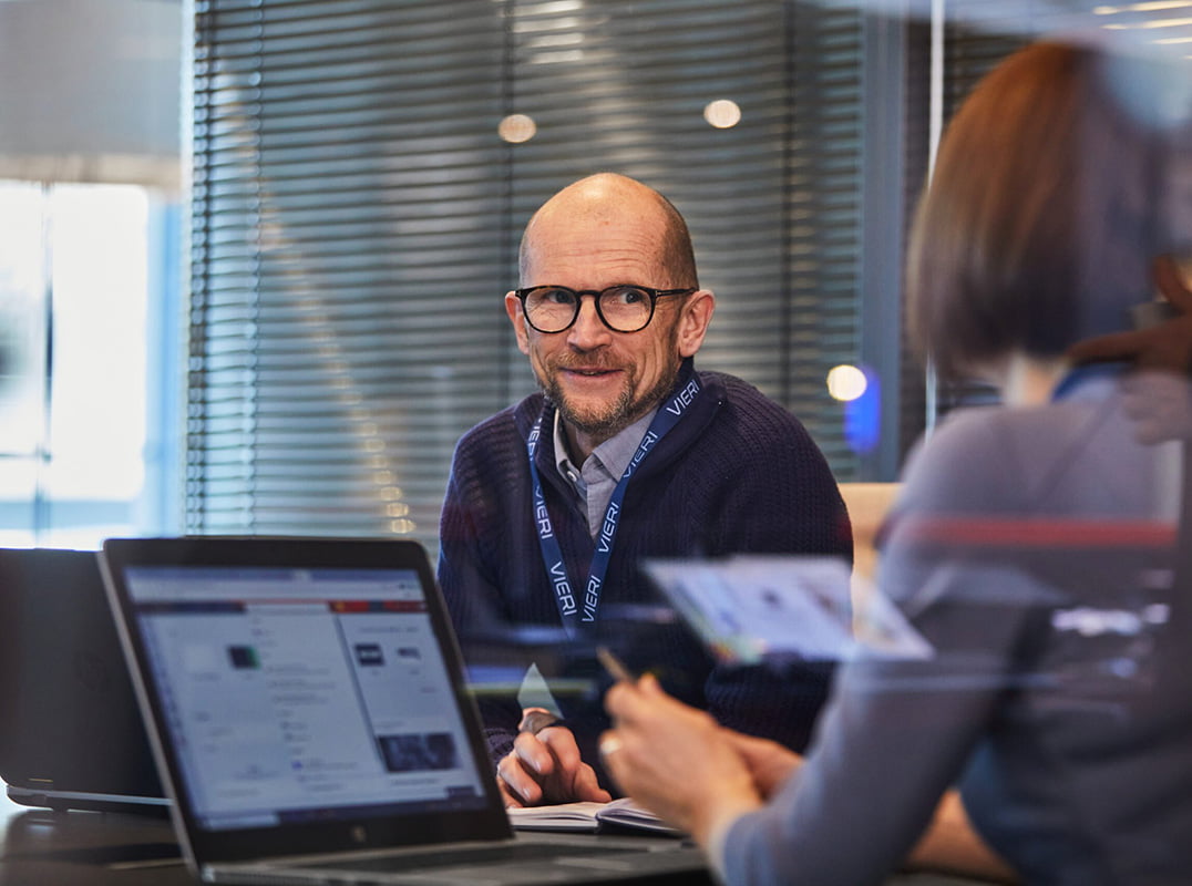 Mann med briller, nøkkelbånd, ser på dame som sitter med ryggen til, foran bærbar PC.