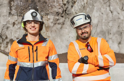 To menn i oransje arbeidsdresser på en byggeplass, med hvite hjelmer