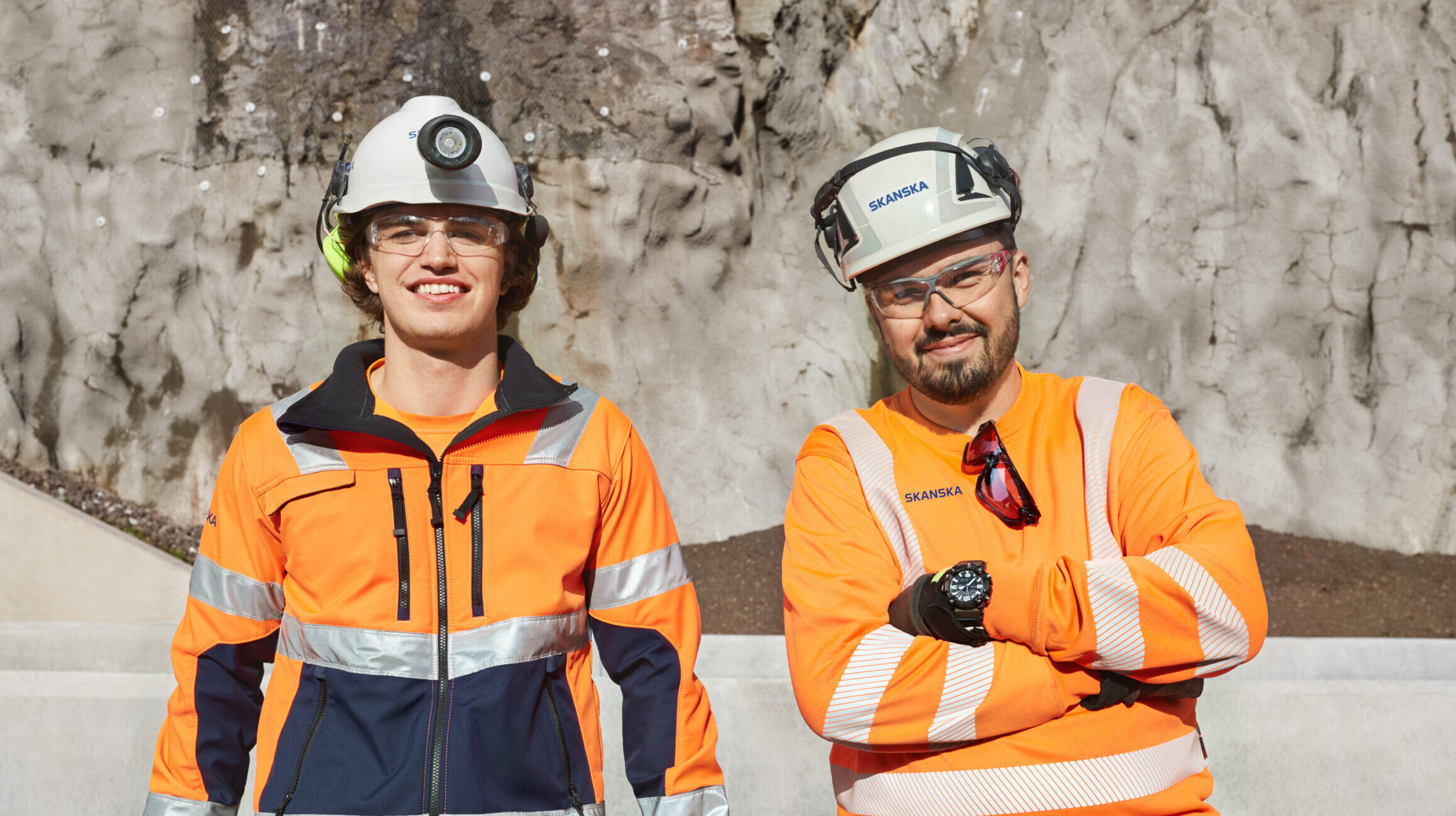 To menn i oransje arbeidsdresser på en byggeplass, med hvite hjelmer